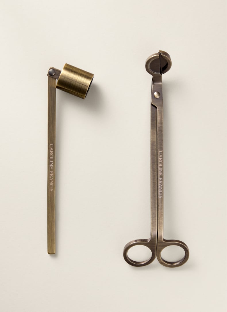 A pair of brass candle tongs and a brass wick holder, both engraved with the brand name 'CAROLINE FRANCIS', displayed on a clean, light beige background.