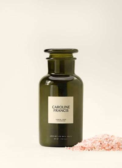 An amber glass bath salts bottle with a cream label reading 'CAROLINE FRANCIS' and 'JAMAICAN ROSE CLEMMONWOOD', shown on a warm off-white background with pink bath salts in the foreground.