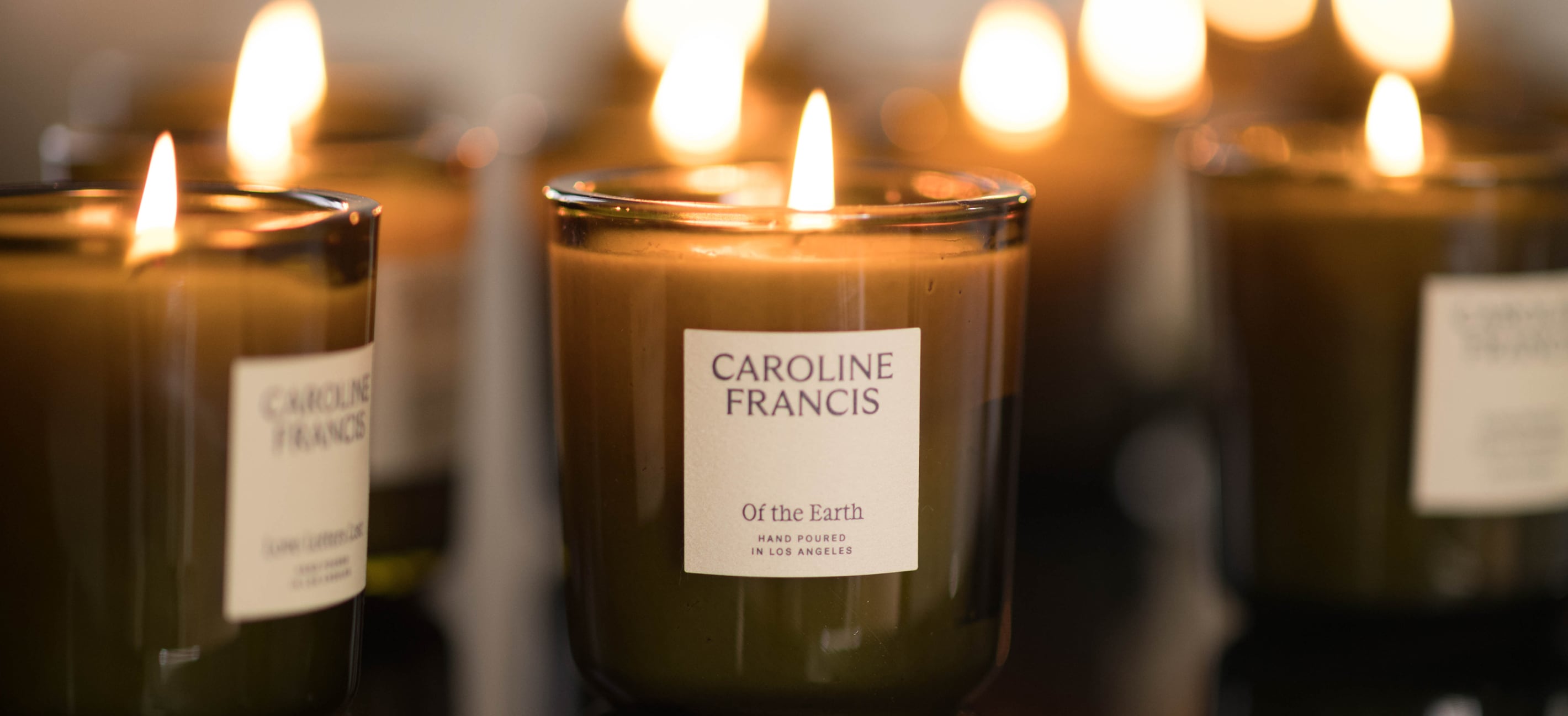 A lit Caroline Francis candle in a clear glass jar with a white label reading 'Of the Earth', surrounded by other blurred candles with warm glowing flames.